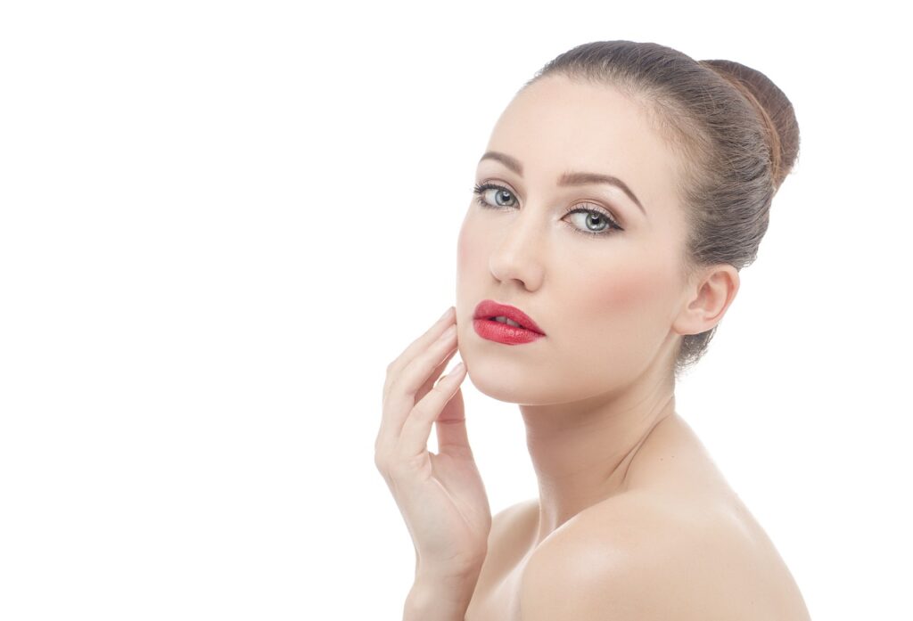 A close-up of a woman with flawless skin and vibrant red lips, gently touching her face.