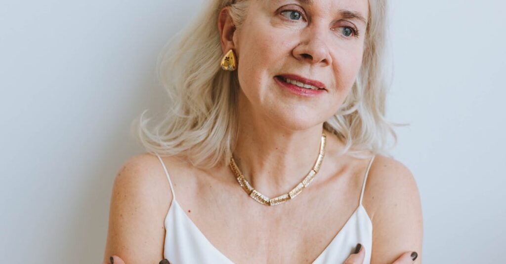 An older woman with light hair, wearing a white top and gold jewelry, sitting calmly with arms crossed, showcasing her radiant but delicate skin.