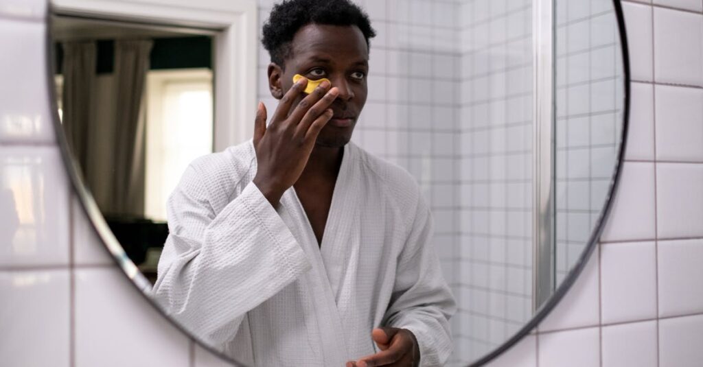 A man in a white robe applying skincare cream under his eyes in front of a bathroom mirror.