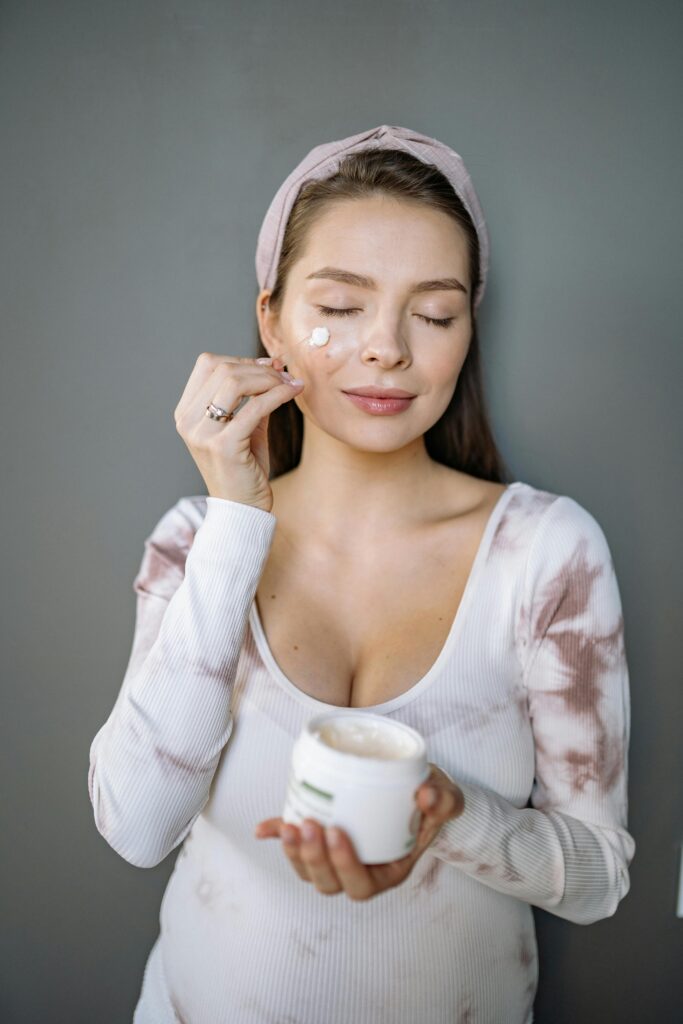 A woman applying cream to her face, holding a jar of moisturizer, preparing her skin for makeup application.