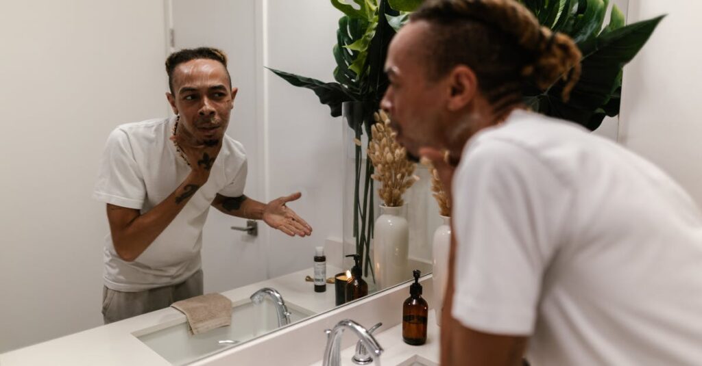 A man cleansing his face in front of a bathroom mirror, wearing a white t-shirt and applying skincare products.