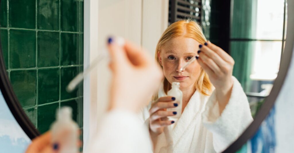 Adult woman applying skincare serum while wearing a bathrobe in front of a bathroom mirror.