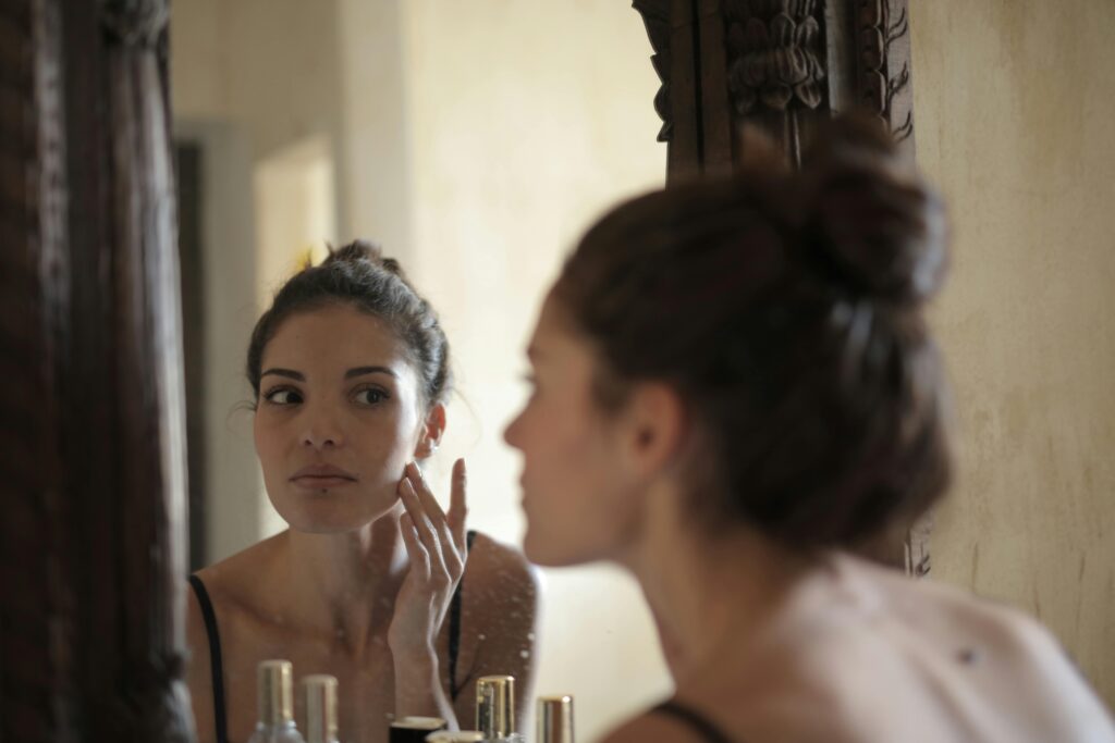 A woman examining her skin in the mirror while touching her face, reflecting on her skincare routine.