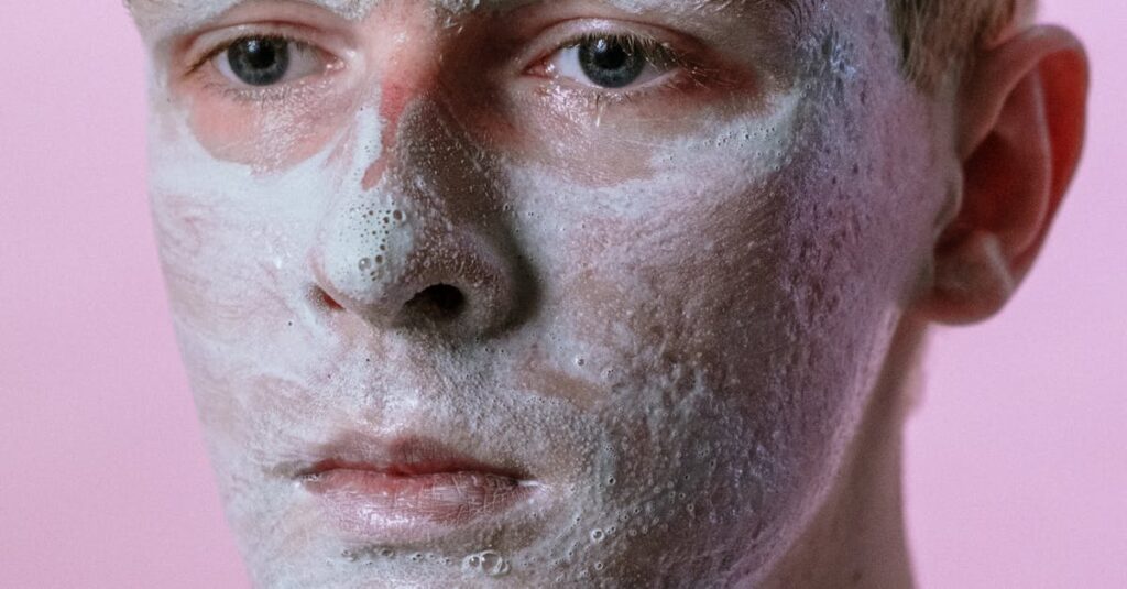 A man with a clay mask on his face against a pastel pink backdrop.