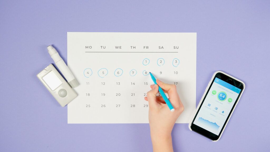 Hand marking a calendar with a blue pen, surrounded by a glucose monitor, a pen-like device, and a smartphone displaying health data.