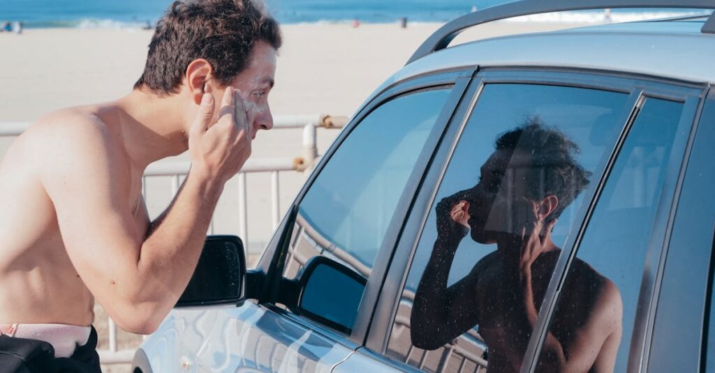 A man applies sunscreen using his car window reflection at a sunny beach.